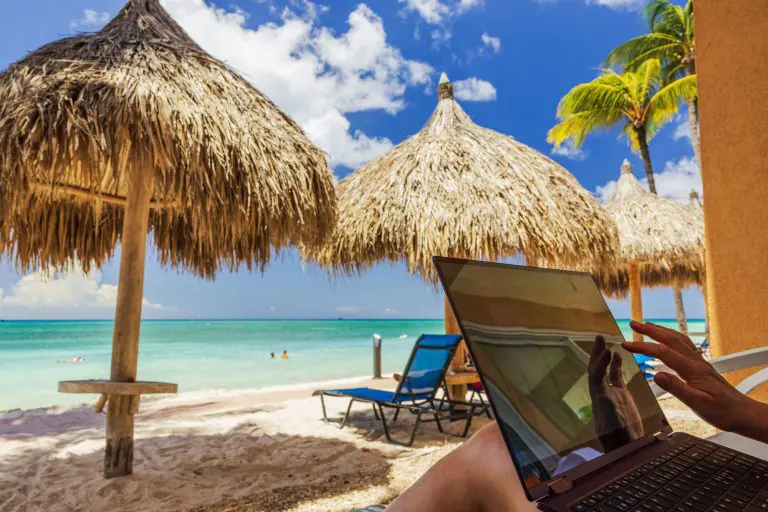 Cómo encontrar mejores precios de alojamiento para una escapada o vacaciones Persona trabajando con un portátil frente a una playa tropical durante unas vacaciones.